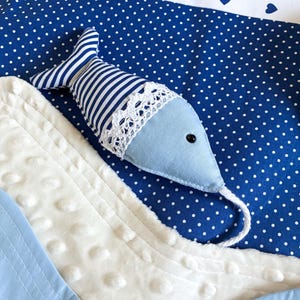 May include: A blue and white nautical-themed baby blanket with a stuffed fish. The fish has a light blue body, striped tail, and a white lace trim. The blanket features a blue polka dot pattern, white fabric, and blue heart accents.