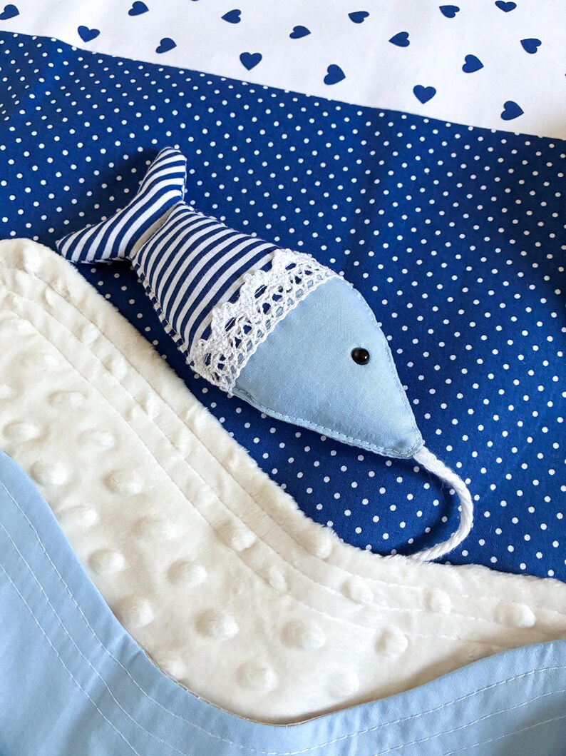 May include: A blue and white nautical-themed baby blanket with a stuffed fish. The fish has a light blue body, striped tail, and a white lace trim. The blanket features a blue polka dot pattern, white fabric, and blue heart accents.