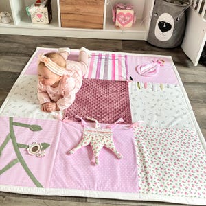 May include: A pink and white baby play mat with various textures and patterns. The mat features a quilted design with floral, polka dot, and striped sections. A star-shaped toy and other sensory elements are attached. A baby is lying on the mat.