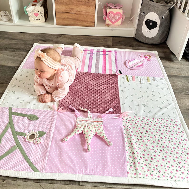 May include: A pink and white baby play mat with various textures and patterns. The mat features a quilted design with floral, polka dot, and striped sections. A star-shaped toy and other sensory elements are attached. A baby is lying on the mat.