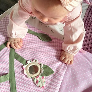 May include: A baby in a pink outfit is reaching for a soft play mat. The mat features a pink and white chevron pattern, a flower with a mirror, and green leaf accents. The mat is designed for infant play and development.