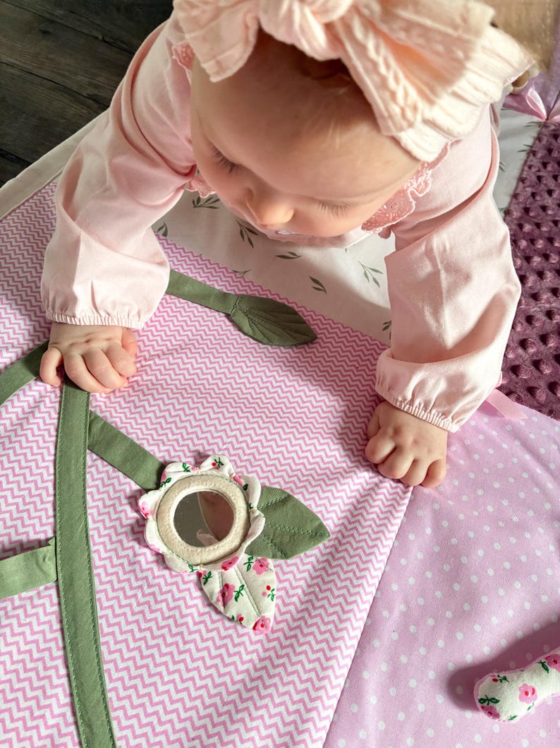 May include: A baby in a pink outfit is reaching for a soft play mat. The mat features a pink and white chevron pattern, a flower with a mirror, and green leaf accents. The mat is designed for infant play and development.