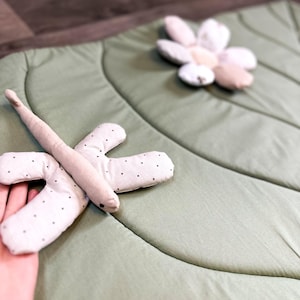 May include: A light green, leaf-shaped play mat with a dragonfly and flower-shaped toys. The dragonfly is beige with black polka dots on its wings. The flower is made of white, beige, and pink petals. The mat is made of soft fabric.