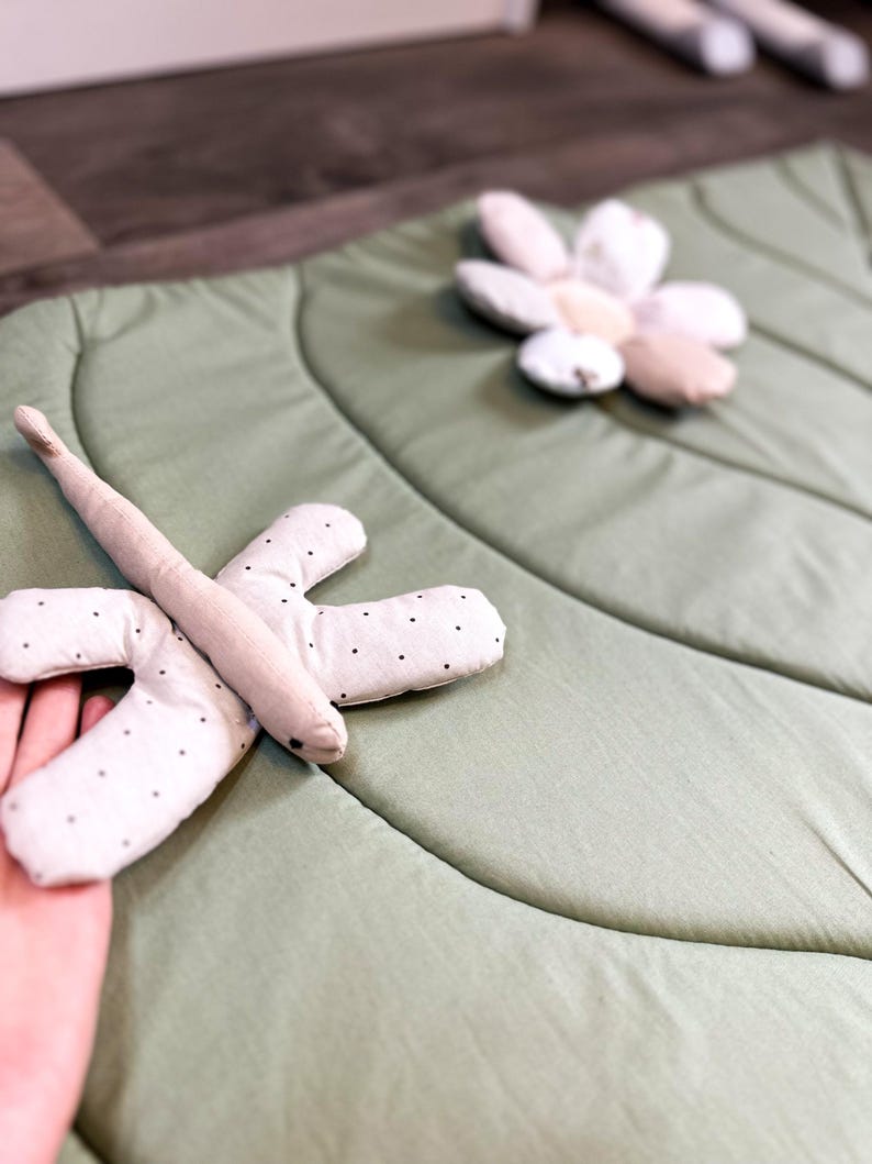 May include: A light green, leaf-shaped play mat with a dragonfly and flower-shaped toys. The dragonfly is beige with black polka dots on its wings. The flower is made of white, beige, and pink petals. The mat is made of soft fabric.