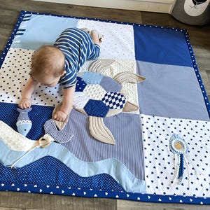 May include: A patchwork baby play mat in shades of blue and white, featuring a turtle design with quilted details. The mat includes a wavy border and a small toy. The mat is designed for babies.