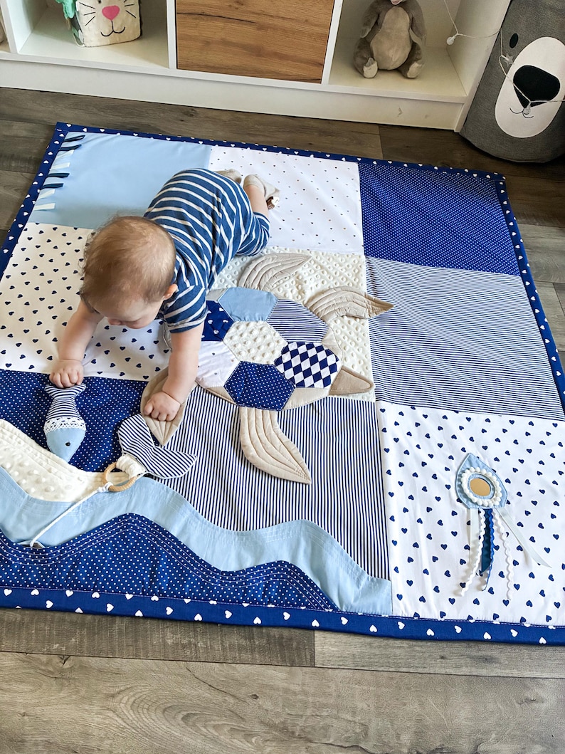 May include: A patchwork baby play mat in shades of blue and white, featuring a turtle design with quilted details. The mat includes a wavy border and a small toy. The mat is designed for babies.