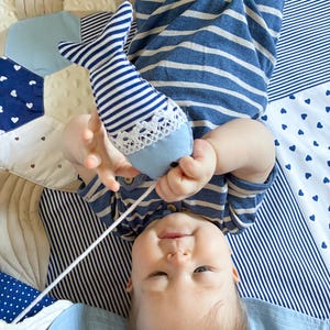 May include: A baby in a blue and white striped outfit holds a blue and white striped fish-shaped toy with a white lace trim. The baby is lying on a patchwork quilt with blue and white patterns.