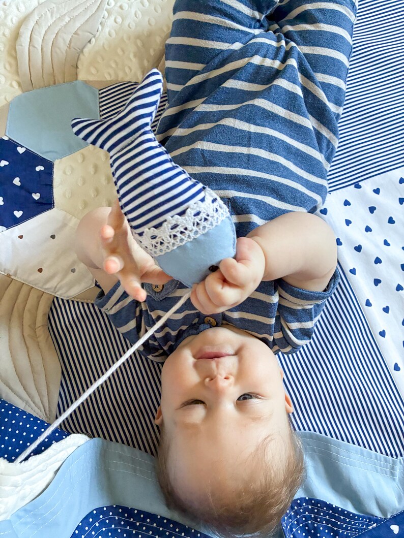 May include: A baby in a blue and white striped outfit holds a blue and white striped fish-shaped toy with a white lace trim. The baby is lying on a patchwork quilt with blue and white patterns.