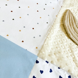 May include: A patchwork baby blanket with various patterns and textures. The blanket features sections of white fabric with small brown, gray, and gold heart-shaped dots, a light blue square, and a section with navy blue hearts. A cream-colored, textured section is also visible.