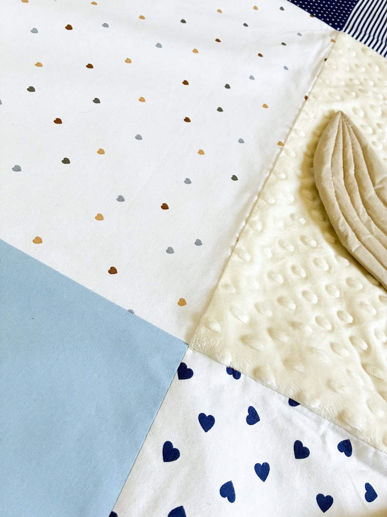 May include: A patchwork baby blanket with various patterns and textures. The blanket features sections of white fabric with small brown, gray, and gold heart-shaped dots, a light blue square, and a section with navy blue hearts. A cream-colored, textured section is also visible.
