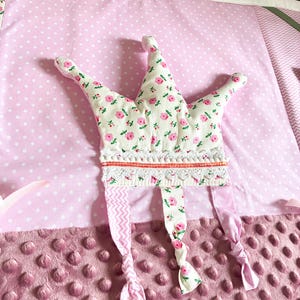 May include: A soft, pink baby play mat with a floral crown. The crown is white with pink flowers and has a lace trim. The mat has a polka dot pattern and a textured, dimpled surface.