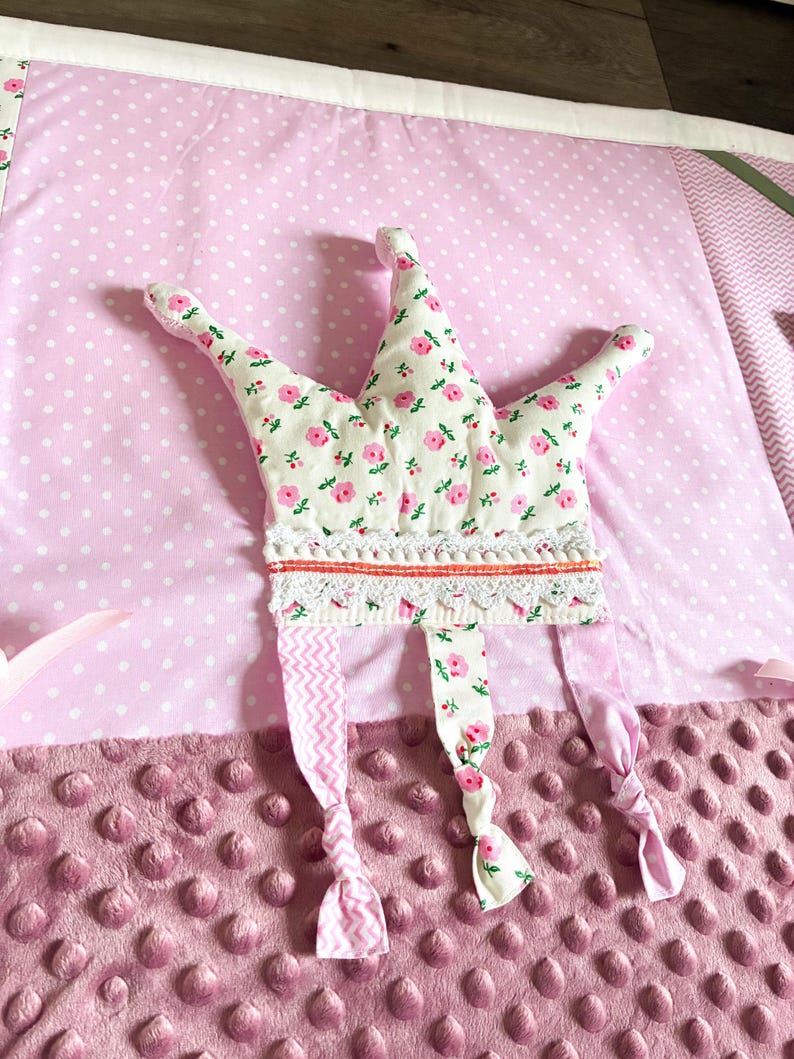 May include: A soft, pink baby play mat with a floral crown. The crown is white with pink flowers and has a lace trim. The mat has a polka dot pattern and a textured, dimpled surface.