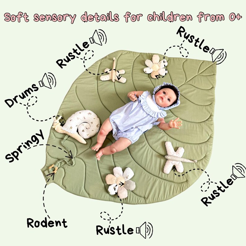 May include: A baby lies on a large, leaf-shaped play mat. The mat is green with textured details and has several attached sensory toys, including flowers, a snail, and a dragonfly. The text "Soft sensory details for children from 0+" is at the top.