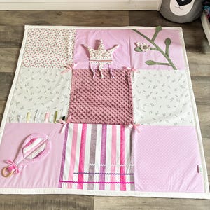 May include: A pink and white patchwork play mat with various textures and patterns. Features include a crown, a rattle, and a striped section. The mat is designed with a baby or toddler in mind.
