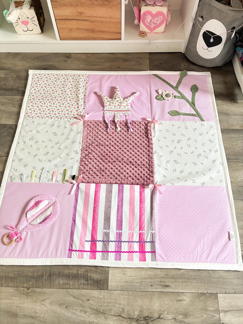 May include: A pink and white patchwork play mat with various textures and patterns. Features include a crown, a rattle, and a striped section. The mat is designed with a baby or toddler in mind.