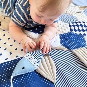 May include: A baby in a blue and white striped shirt is reaching for a blue and white striped fish-shaped toy on a patchwork play mat. The mat features various blue and white patterns, including polka dots and hearts.