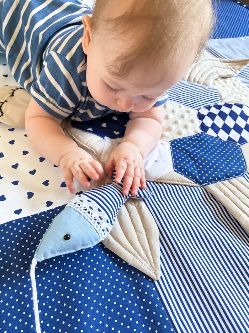 May include: A baby in a blue and white striped shirt is reaching for a blue and white striped fish-shaped toy on a patchwork play mat. The mat features various blue and white patterns, including polka dots and hearts.
