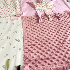 May include: A patchwork baby play mat with various fabrics. Features include a pink dimpled fabric, floral print, and a star-shaped toy with floral accents. Pink ribbons and green leaves add detail.