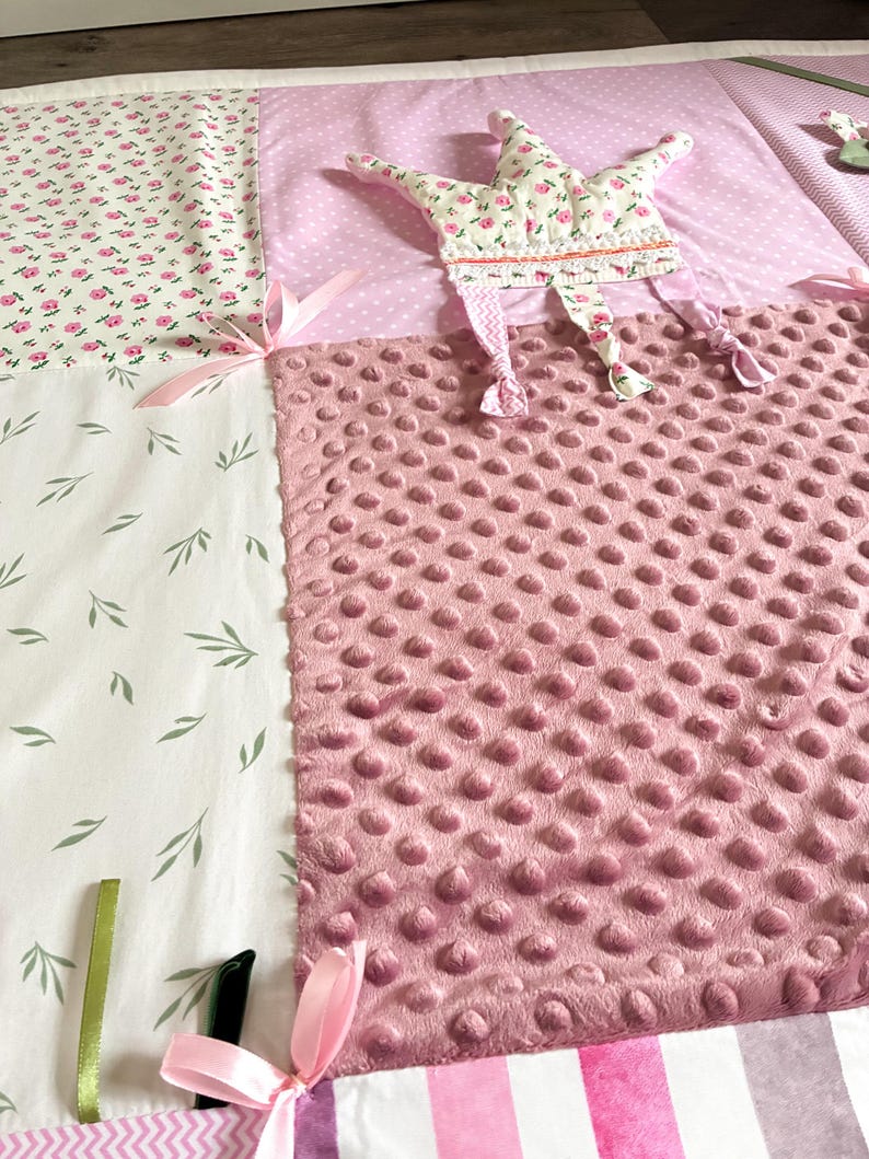 May include: A patchwork baby play mat with various fabrics. Features include a pink dimpled fabric, floral print, and a star-shaped toy with floral accents. Pink ribbons and green leaves add detail.