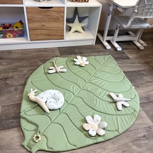 May include: A light green, leaf-shaped play mat with quilted details. The mat is decorated with plush, whimsical accents, including a snail, flowers, and dragonflies. The mat is designed for babies and toddlers.