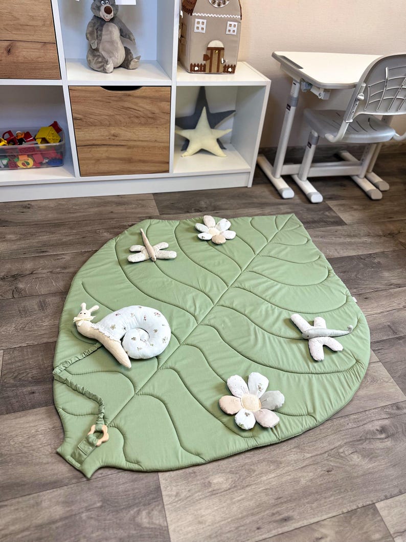 May include: A light green, leaf-shaped play mat with quilted details. The mat is decorated with plush, whimsical accents, including a snail, flowers, and dragonflies. The mat is designed for babies and toddlers.
