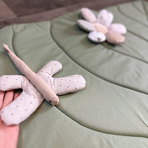May include: A light green leaf-shaped play mat with a soft, quilted texture. A beige dragonfly toy with black polka dots is on the mat. A flower-shaped pillow in shades of beige and white is also on the mat. The mat is designed for babies and toddlers.