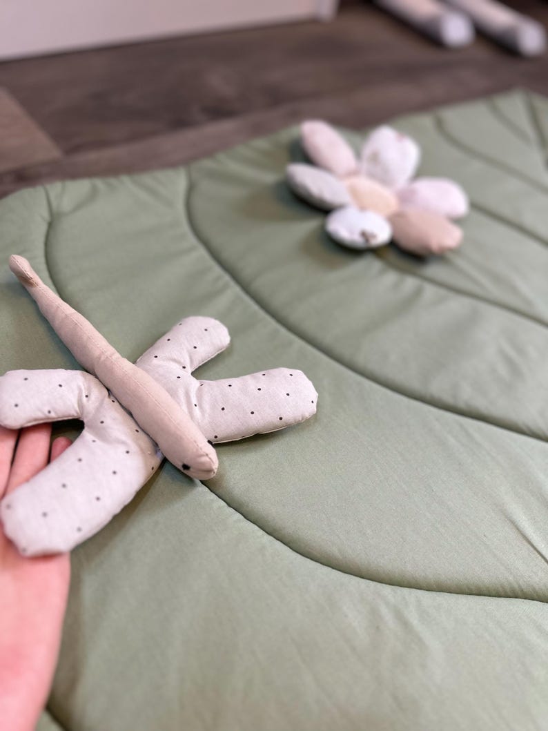 May include: A light green leaf-shaped play mat with a soft, quilted texture. A beige dragonfly toy with black polka dots is on the mat. A flower-shaped pillow in shades of beige and white is also on the mat. The mat is designed for babies and toddlers.