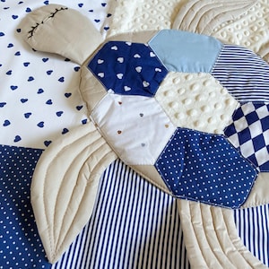 May include: A quilted turtle-shaped blanket with a patchwork design. The turtle features a beige head and flippers, and a shell made of blue and white patterned fabrics, including hearts, polka dots, and stripes. The background is a mix of white and blue fabrics.