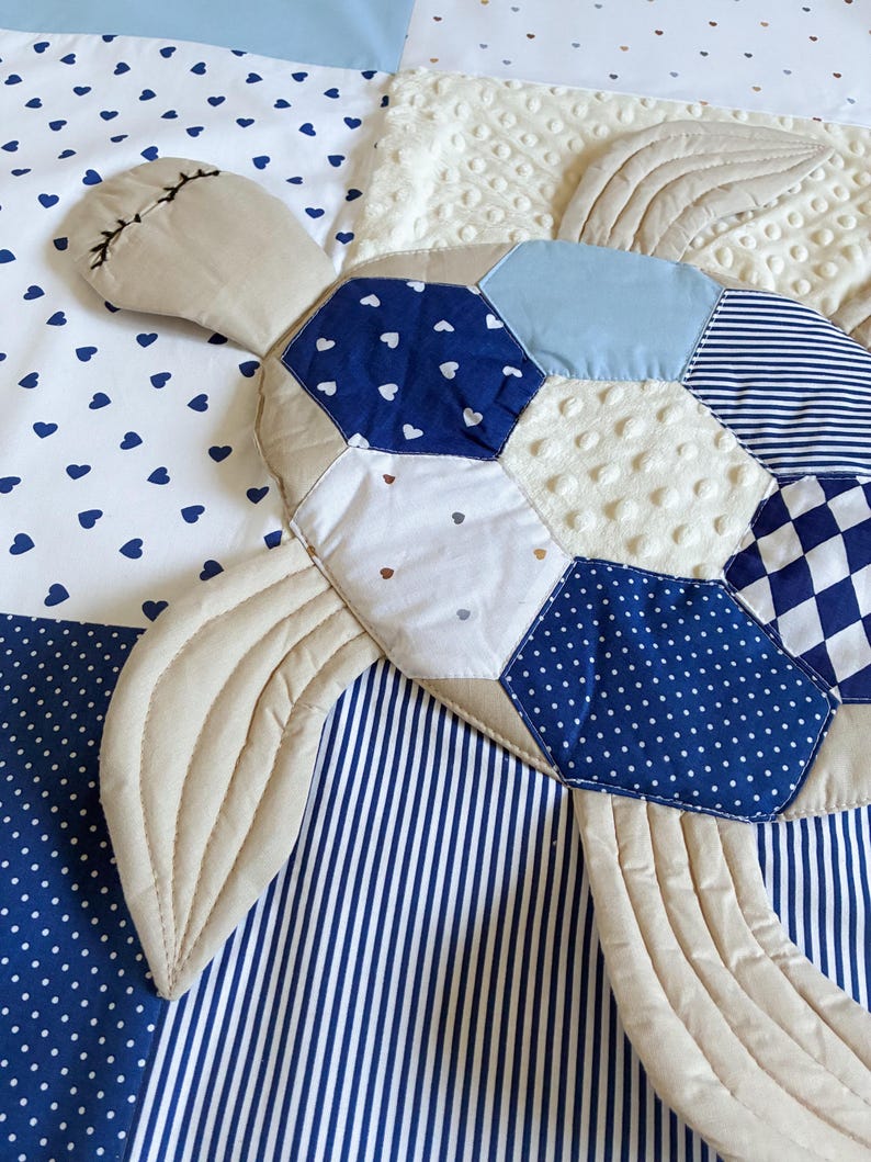 May include: A quilted turtle-shaped blanket with a patchwork design. The turtle features a beige head and flippers, and a shell made of blue and white patterned fabrics, including hearts, polka dots, and stripes. The background is a mix of white and blue fabrics.