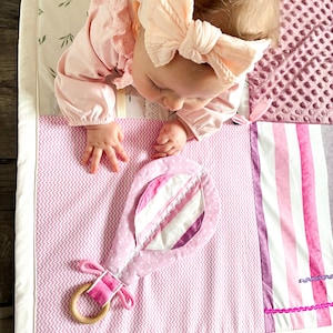 May include: A baby lies on a pink and white patterned play mat with a soft toy. The mat features a variety of textures and patterns, including stripes, polka dots, and a textured plush section. The toy is a pink rattle with a wooden ring.