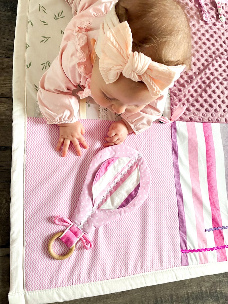 May include: A baby lies on a pink and white patterned play mat with a soft toy. The mat features a variety of textures and patterns, including stripes, polka dots, and a textured plush section. The toy is a pink rattle with a wooden ring.