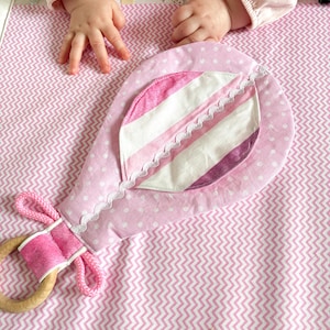 May include: A pink baby rattle with a wooden ring and a fabric design. The rattle features a polka dot pattern and a striped accent, with a scalloped lace trim. The handle is wrapped in pink fabric and rope.