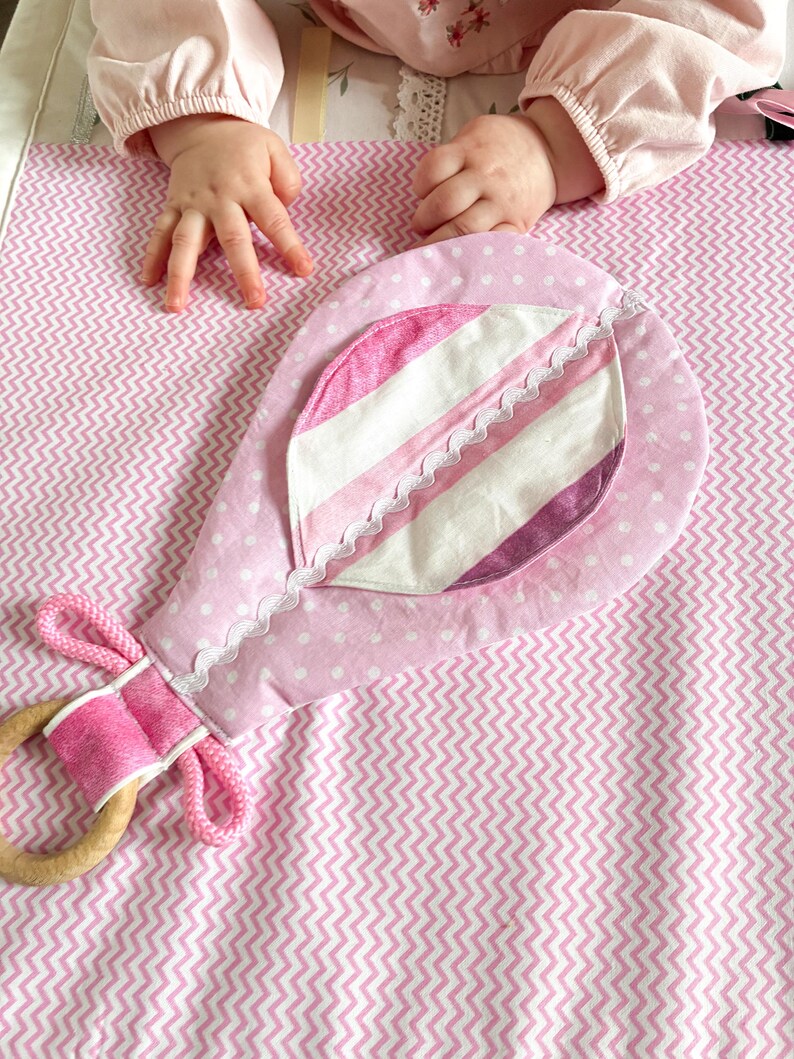 May include: A pink baby rattle with a wooden ring and a fabric design. The rattle features a polka dot pattern and a striped accent, with a scalloped lace trim. The handle is wrapped in pink fabric and rope.