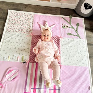 May include: A baby wearing a crown and pink outfit lies on a patchwork play mat. The mat features various pink patterns, including floral prints, stripes, and polka dots. A toy mirror and decorative elements are attached to the mat.