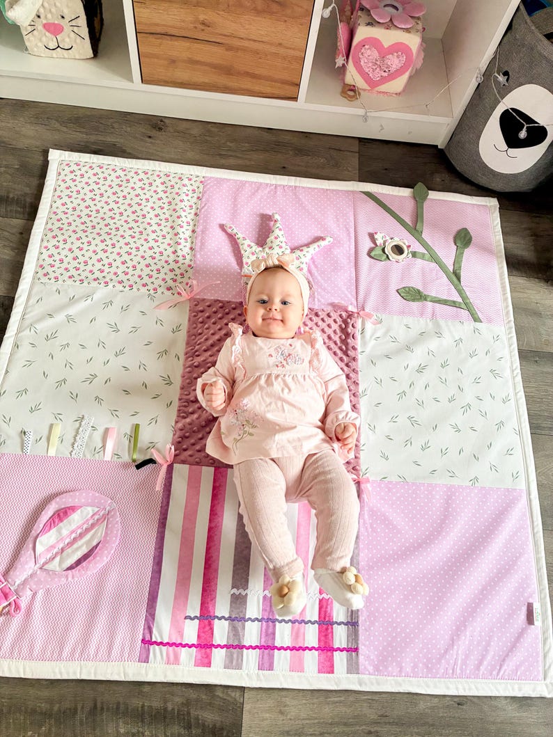 May include: A baby wearing a crown and pink outfit lies on a patchwork play mat. The mat features various pink patterns, including floral prints, stripes, and polka dots. A toy mirror and decorative elements are attached to the mat.