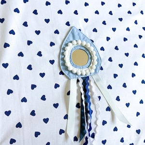 May include: A white fabric with a navy blue heart pattern. A light blue teardrop-shaped applique with a small mirror, white pom-poms, and ribbons in white, navy, and dark blue. The top edge has navy blue stripes.