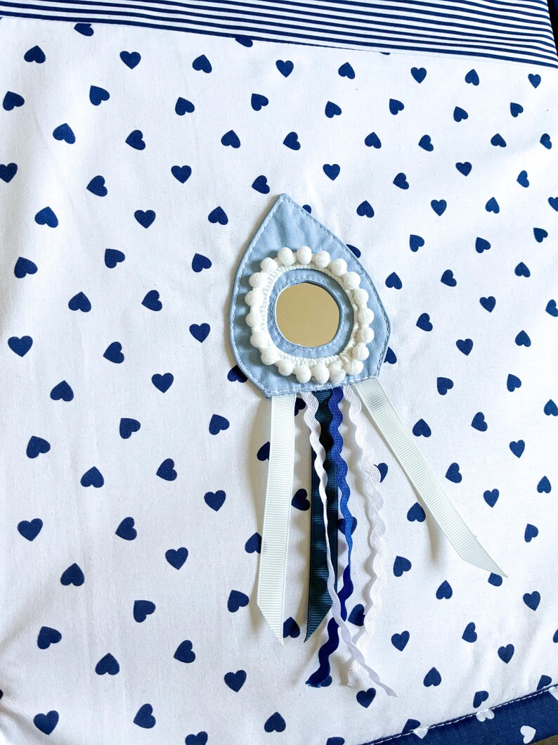 May include: A white fabric with a navy blue heart pattern. A light blue teardrop-shaped applique with a small mirror, white pom-poms, and ribbons in white, navy, and dark blue. The top edge has navy blue stripes.