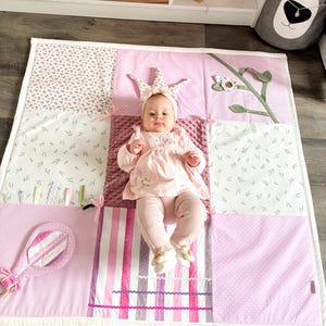 May include: A baby wearing a crown headband and pink outfit lies on a patchwork play mat. The mat features various pink patterns, including floral, polka dots, and stripes. A pink rattle is on the left side of the mat.
