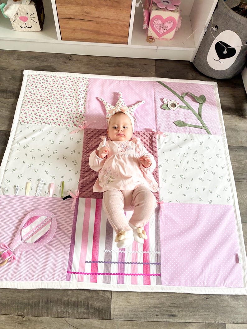 May include: A baby wearing a crown headband and pink outfit lies on a patchwork play mat. The mat features various pink patterns, including floral, polka dots, and stripes. A pink rattle is on the left side of the mat.