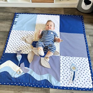 May include: A baby lies on a blue and white patchwork play mat with a whale tail design. The mat features various patterns, including polka dots, stripes, and solid colors. The baby wears a blue and white striped outfit.