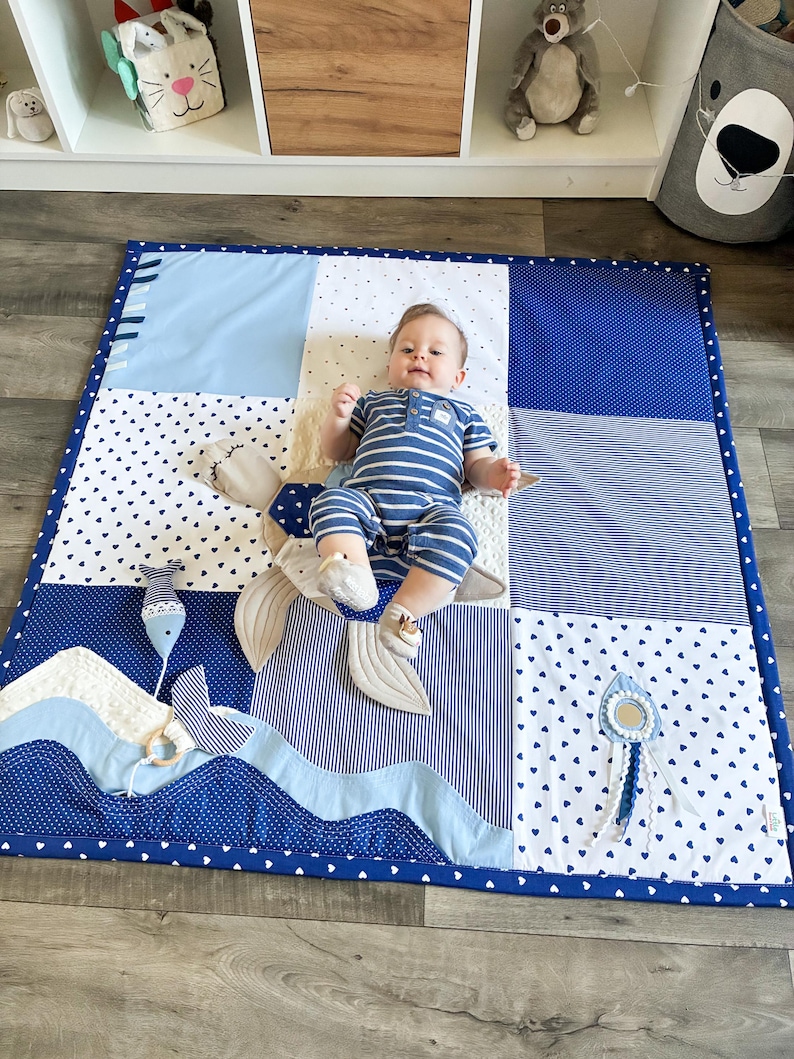 May include: A baby lies on a blue and white patchwork play mat with a whale tail design. The mat features various patterns, including polka dots, stripes, and solid colors. The baby wears a blue and white striped outfit.