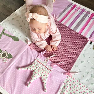 May include: A patchwork baby play mat in shades of pink, white, and green. The mat features various patterns, including polka dots, floral prints, and stripes. A soft, plush toy with a floral pattern is on the mat.