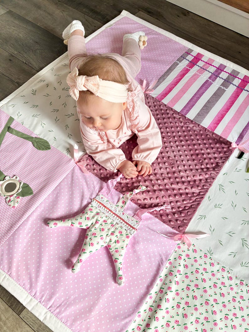 May include: A patchwork baby play mat in shades of pink, white, and green. The mat features various patterns, including polka dots, floral prints, and stripes. A soft, plush toy with a floral pattern is on the mat.