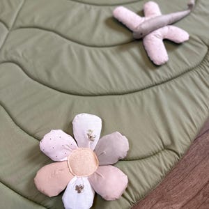 May include: A soft, sage green leaf-shaped play mat with a quilted texture. A plush flower and dragonfly toy rest on the mat. The flower has pink, beige, and white petals with embroidered details. The dragonfly is light gray.