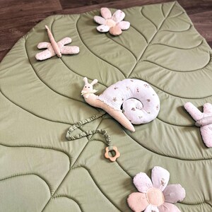 May include: A light green leaf-shaped play mat with quilted details. The mat is decorated with plush toys, including a snail, dragonfly, and several flowers. The snail has a cream-colored shell with floral patterns and a wooden teething ring.