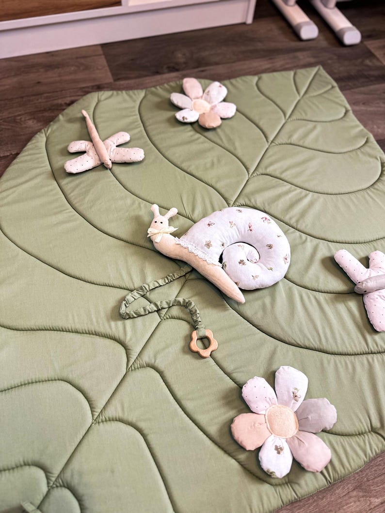 May include: A light green leaf-shaped play mat with quilted details. The mat is decorated with plush toys, including a snail, dragonfly, and several flowers. The snail has a cream-colored shell with floral patterns and a wooden teething ring.