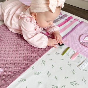 May include: A baby wearing a pink outfit and a matching bow headband is lying on a soft, multi-textured play mat. The mat features a variety of patterns, including a dotted fabric, stripes, and a white fabric with green leaf designs. Several colorful clips are on the mat.