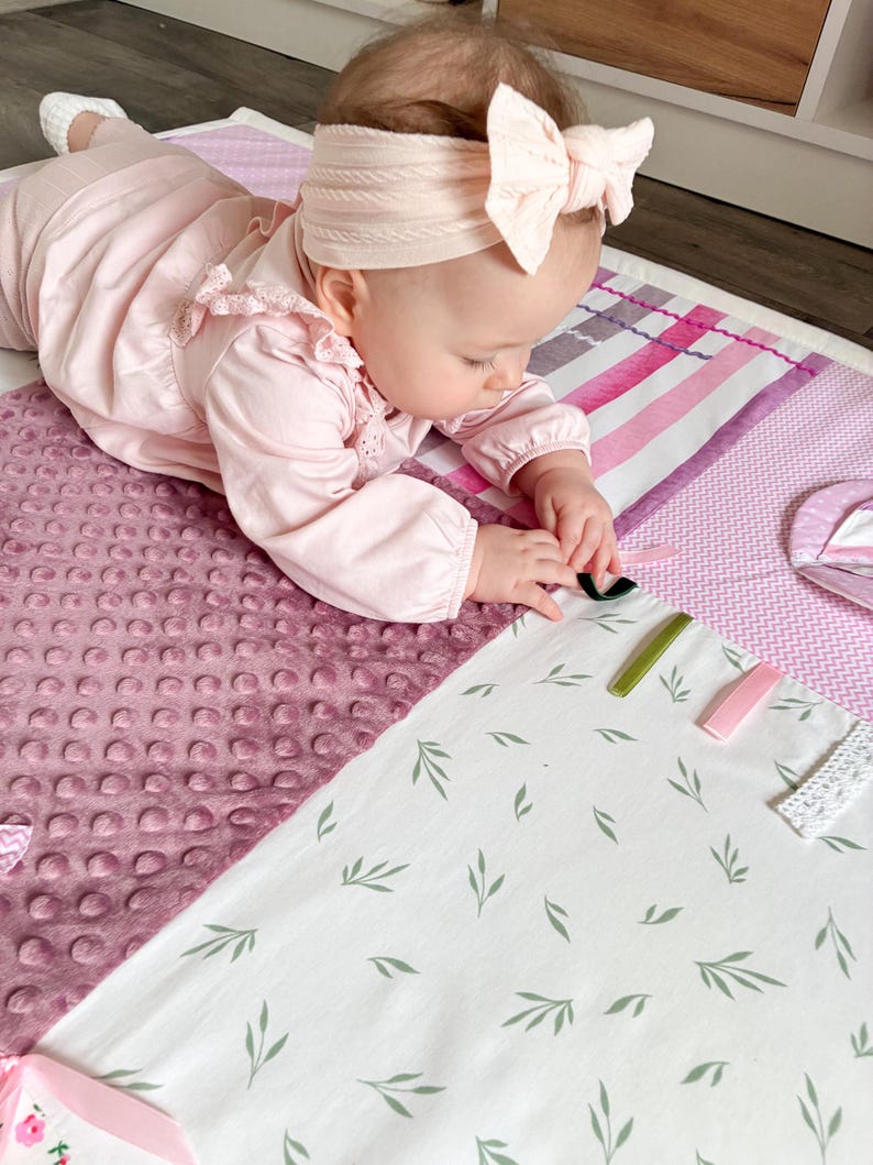 May include: A baby wearing a pink outfit and a matching bow headband is lying on a soft, multi-textured play mat. The mat features a variety of patterns, including a dotted fabric, stripes, and a white fabric with green leaf designs. Several colorful clips are on the mat.