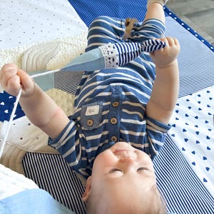 May include: A baby wearing a blue and white striped onesie lies on a patchwork play mat. The mat features various blue and white patterns, including polka dots and stripes. The baby is holding a blue and white toy with a white lace trim.