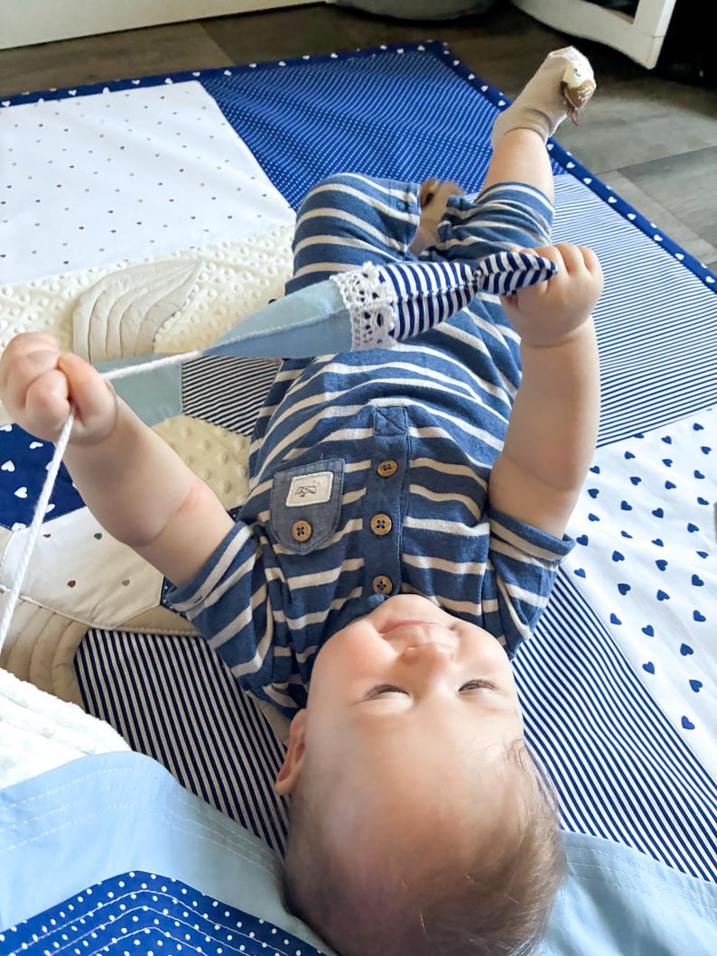 May include: A baby wearing a blue and white striped onesie lies on a patchwork play mat. The mat features various blue and white patterns, including polka dots and stripes. The baby is holding a blue and white toy with a white lace trim.